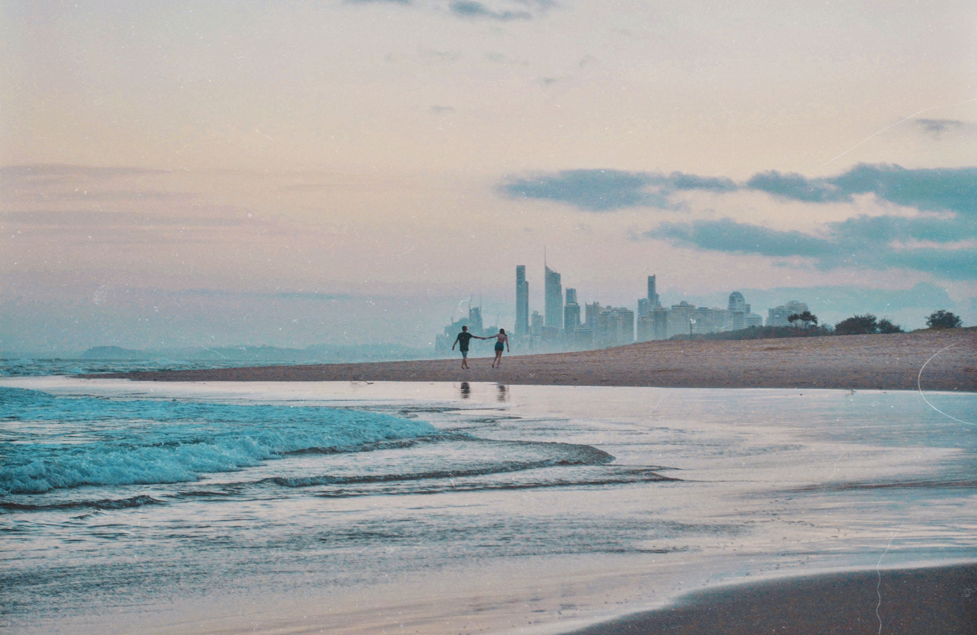 Twee mensen lopen hand in hand over het strand in Australië, met op de achtergrond een stad gelegen en op de voorgrond de zee, met ondergaande zon