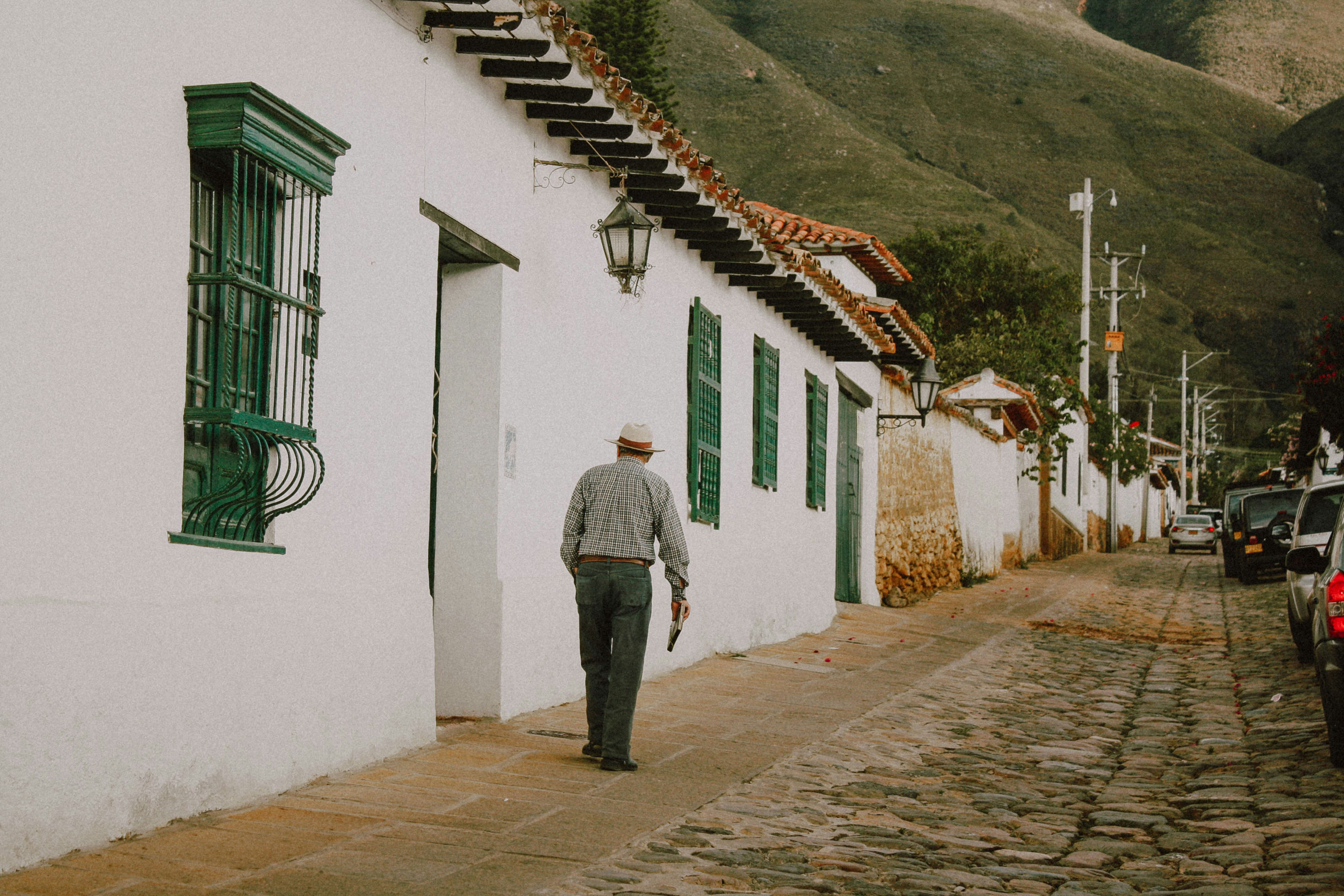 Old man walking past white plastered houses down a cobbled street in Villa de Leyva, with green hills in the background